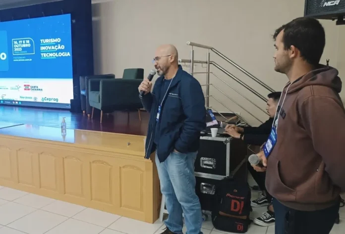 AMESC sedia 2º Fórum de Turismo Caminho dos Canyons Palestrante com microfone, em pé no palco, em frente ao painel do "Fórum de Turismo - Caminho dos Canyons", evento da AMESC realizado em Araranguá para debater a inovação no setor.