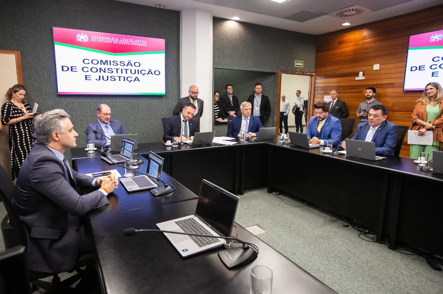 Mesa de reuniões da Comissão de Constituição e Justiça (CCJ) da Assembleia Legislativa de Santa Catarina (Alesc) com deputados e assessores sentados e o painel eletrônico ao fundo.