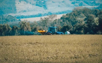 Alesc reúne sete comissões para votar 13 PLs do Executivo Alesc reúne sete comissões para votar 13 PLs do Executivo.webp: Trator azul trabalhando em um campo de colheita amarelado ou bege em primeiro plano. O trator possui um implemento acoplado que espalha fertilizante ou sementes. O fundo mostra uma paisagem montanhosa e arborizada, com outras propriedades e morros em diferentes tons de verde e azul, sugerindo uma área rural.