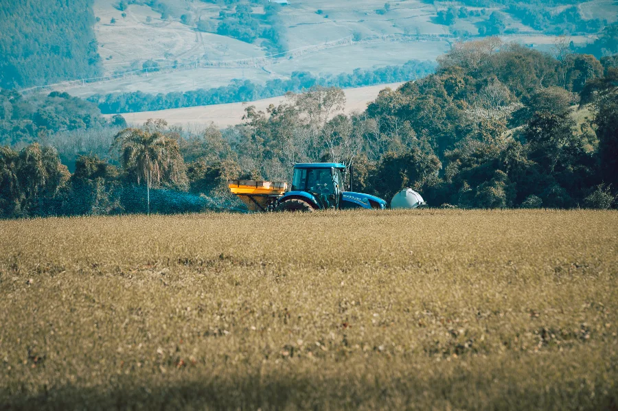 Alesc reúne sete comissões para votar 13 PLs do Executivo.webp: Trator azul trabalhando em um campo de colheita amarelado ou bege em primeiro plano. O trator possui um implemento acoplado que espalha fertilizante ou sementes. O fundo mostra uma paisagem montanhosa e arborizada, com outras propriedades e morros em diferentes tons de verde e azul, sugerindo uma área rural.