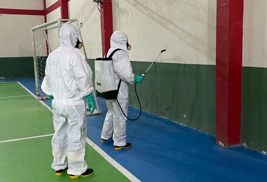 Agentes de saúde em macacões brancos e máscaras, realizando a Borrifação Residual Intradomiciliar (BRI) de inseticida em uma quadra esportiva, como parte da intensificação do combate à dengue em Araranguá.