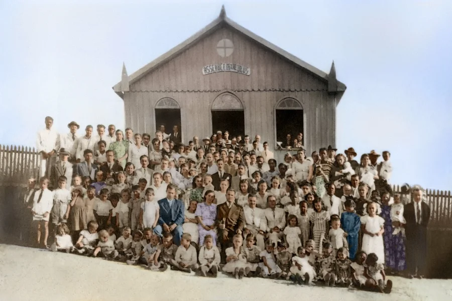 Fotografia antiga (colorida digitalmente) da primeira sede de madeira da Assembleia de Deus de Criciúma, com um grande grupo de fiéis (homens, mulheres e crianças) reunidos na frente, ilustrando o início da história da igreja.