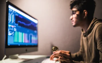 BC cria marco regulatório para o mercado de criptoativos Pessoa segurando uma moeda de Bitcoin (criptoativo) em frente a um monitor que exibe gráficos de negociação, ilustrando o mercado financeiro e o novo marco regulatório do Banco Central (BC) para as Sociedades Prestadoras de Serviços de Ativos Virtuais (SPSVAs) no Brasil.