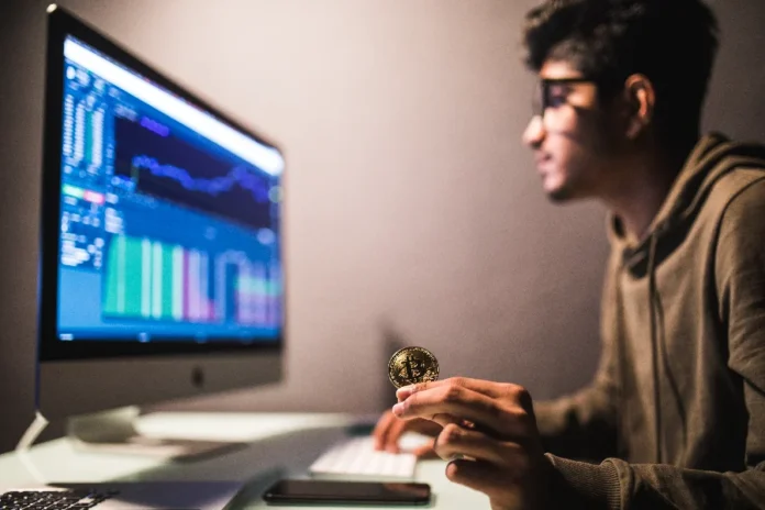 BC cria marco regulatório para o mercado de criptoativos Pessoa segurando uma moeda de Bitcoin (criptoativo) em frente a um monitor que exibe gráficos de negociação, ilustrando o mercado financeiro e o novo marco regulatório do Banco Central (BC) para as Sociedades Prestadoras de Serviços de Ativos Virtuais (SPSVAs) no Brasil.