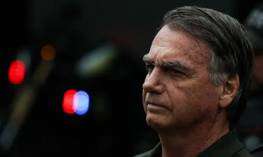 Bolsonaro é preso pela PF em cumprimento a mandado do STF Ex-presidente Jair Bolsonaro com expressão séria; luzes de veículos policiais, embaçadas, aparecem no fundo.