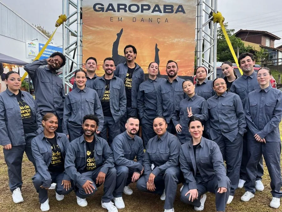 Grupo de bailarinos da Cia de Dança Unesc, vestindo uniformes cinzas, posando juntos em frente a um banner do evento 