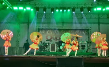 Ciclone: Criciúma retoma Quermesse às 17h deste sábado Dançarinas com fantasias coloridas e sombrinhas se apresentando no palco iluminado da Quermesse 2025 em Criciúma, evento retomado após o alerta de ciclone.