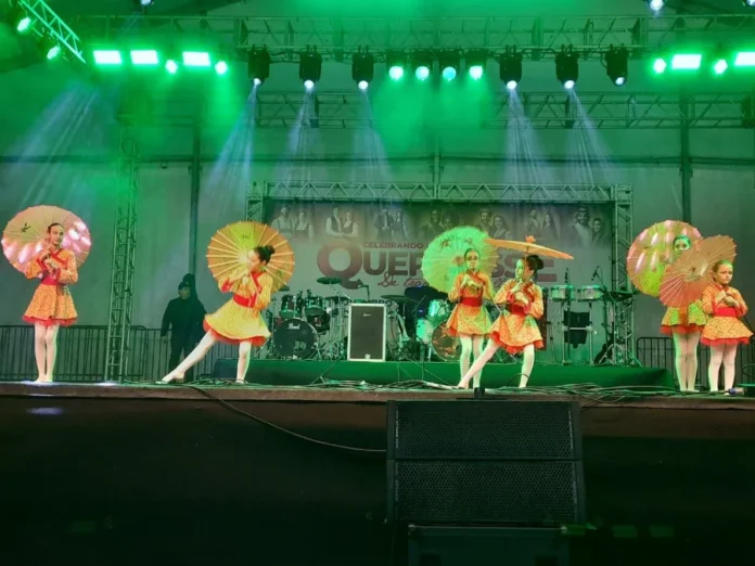 Ciclone Criciúma retoma Quermesse às 17h deste sábado Dançarinas com fantasias coloridas e sombrinhas se apresentando no palco iluminado da Quermesse 2025 em Criciúma, evento retomado após o alerta de ciclone.