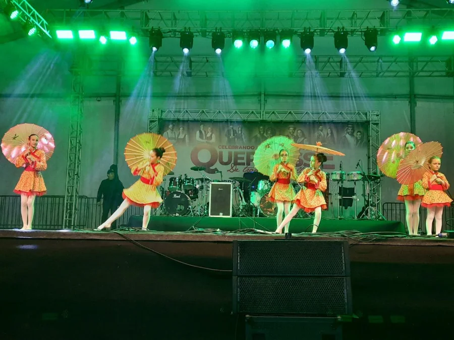 Ciclone Criciúma retoma Quermesse às 17h deste sábado Dançarinas com fantasias coloridas e sombrinhas se apresentando no palco iluminado da Quermesse 2025 em Criciúma, evento retomado após o alerta de ciclone.