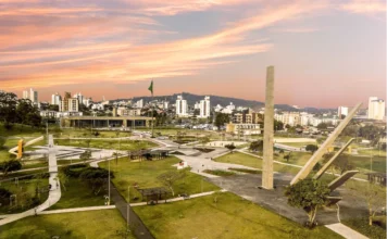 Criciúma: Centenário altera serviços na Saúde e Educação Vista aérea do Paço Municipal Marcos Rovaris e do Parque da Prefeitura em Criciúma, com o obelisco em destaque, ilustrando o feriado do Centenário que altera os serviços municipais de Saúde e Educação.