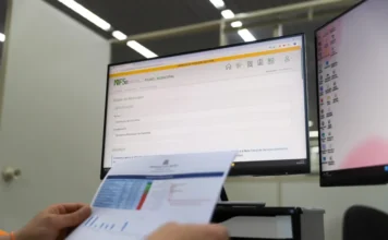 Criciúma adota Emissor Nacional de Nota Fiscal Eletrônica Profissional da Secretaria da Fazenda de Criciúma analisando documentos fiscais em frente a um monitor que exibe o painel de controle do novo sistema de Nota Fiscal de Serviço Eletrônica (NFS-e) Nacional.