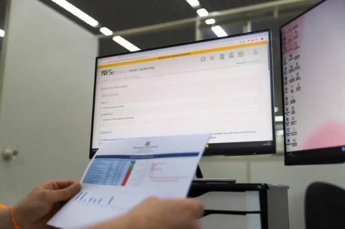 Criciúma adota Emissor Nacional de Nota Fiscal Eletrônica Profissional da Secretaria da Fazenda de Criciúma analisando documentos fiscais em frente a um monitor que exibe o painel de controle do novo sistema de Nota Fiscal de Serviço Eletrônica (NFS-e) Nacional.