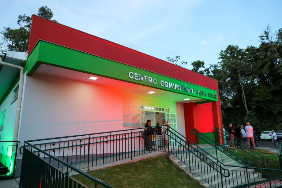 Vista externa e frontal do novo Centro Comunitário Vera Cruz em Criciúma. O prédio tem cores vibrantes em vermelho e verde na fachada superior, com a identificação "Centro Comunitário Vera Cruz" em destaque. Uma rampa de acesso com corrimãos e escadas é visível, com pessoas na entrada e no estacionamento.