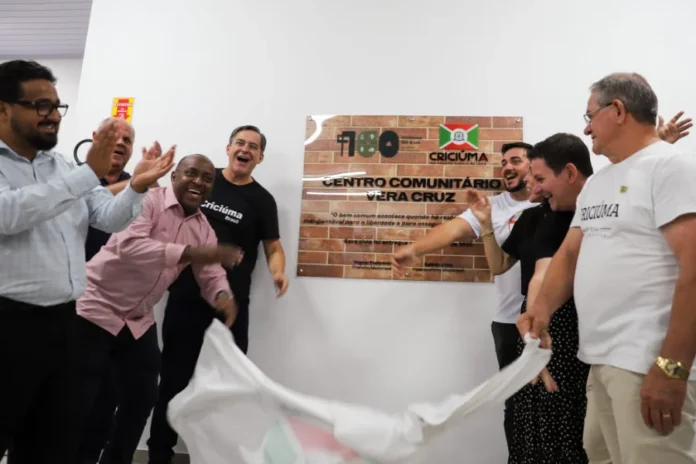 Criciúma inaugura Centro Comunitário no bairro Vera Cruz Autoridades municipais, incluindo o prefeito, vice-prefeito e secretários, sorriem e aplaudem durante a cerimônia de inauguração do Centro Comunitário Vera Cruz. Eles estão descerrando uma placa de tijolos que contém o nome do centro e uma citação.