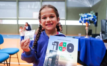 Criciúma lança álbum de figurinhas do Centenário da cidade Criança (aluna da rede municipal) sorrindo e exibindo o álbum de figurinhas "100 Anos de Criciúma", que destaca a história, personagens e pontos turísticos da cidade, em celebração ao Centenário.