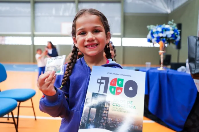 Criciúma lança álbum de figurinhas do Centenário da cidade Criança (aluna da rede municipal) sorrindo e exibindo o álbum de figurinhas "100 Anos de Criciúma", que destaca a história, personagens e pontos turísticos da cidade, em celebração ao Centenário.