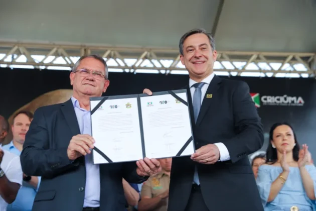 Prefeito Vagner Espindola (à direita) e o vice-prefeito Salésio Lima segurando o documento de autorização para o início do processo licitatório da Cidade do Autista em Criciúma, durante as comemorações do Centenário.
