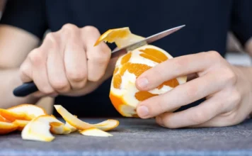 Descascar mais e desembrulhar menos Mãos usando uma faca para descascar uma laranja, ilustrando o conceito nutricional de "descascar mais e desembrulhar menos" (escolher alimentos naturais em vez de industrializados).