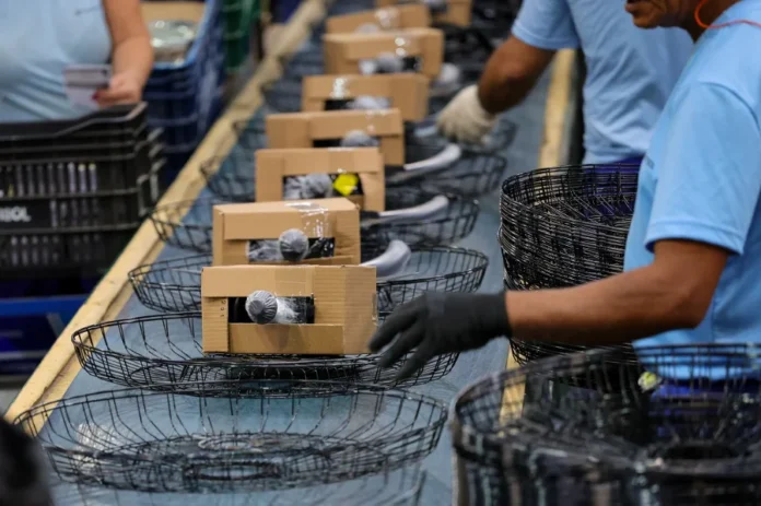 Economia de SC cresce acima da média nacional em 2025 Trabalhadores com luvas em uma linha de montagem industrial, embalando produtos em cestos e caixas, ilustrando o forte crescimento de 3,1% da produção industrial, que impulsiona a economia de Santa Catarina (SC) acima da média nacional.