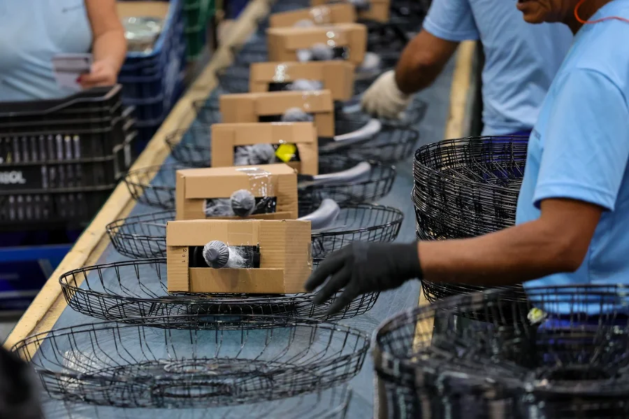 Economia de SC cresce acima da média nacional em 2025 Trabalhadores com luvas em uma linha de montagem industrial, embalando produtos em cestos e caixas, ilustrando o forte crescimento de 3,1% da produção industrial, que impulsiona a economia de Santa Catarina (SC) acima da média nacional.