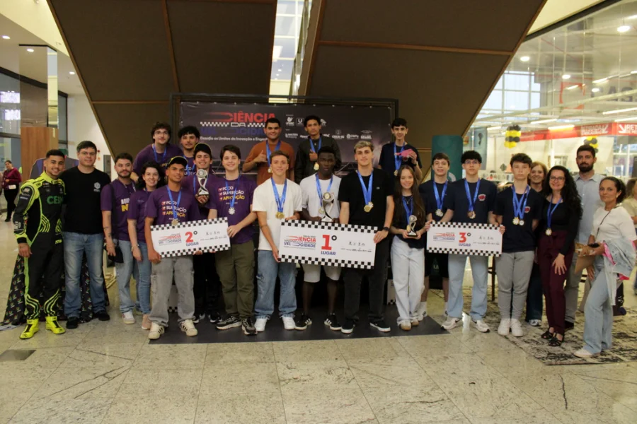 Equipes finalistas do Projeto Ciência da Velocidade posam no pódio, exibindo medalhas e troféus, no Nações Shopping em Criciúma.
