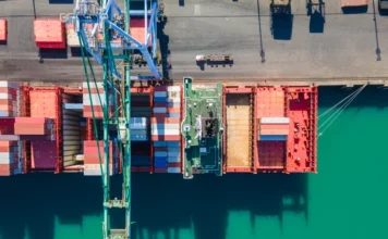 Exportações de SC batem recorde e chegam a US$ 1,1 bi Vista aérea de um navio de contêineres em porto de Santa Catarina, com guindastes descarregando carga sobre o cais, simbolizando o recorde de exportações de US$ 1,1 bilhão em outubro.