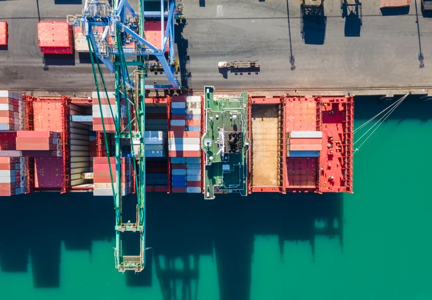 Exportações de SC batem recorde e chegam a US$ 1,1 bi Vista aérea de um navio de contêineres em porto de Santa Catarina, com guindastes descarregando carga sobre o cais, simbolizando o recorde de exportações de US$ 1,1 bilhão em outubro.