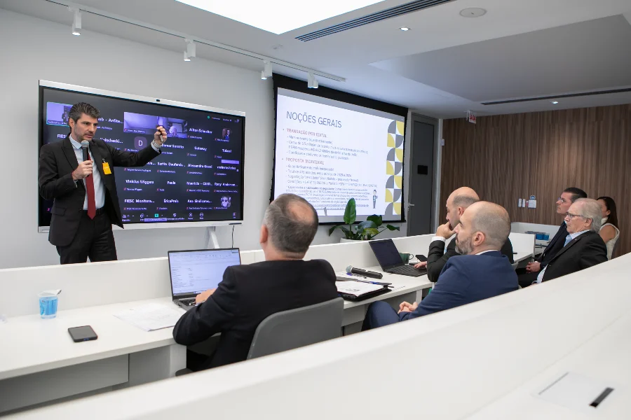 FIESC Nova lei do IR tem lacunas e gera insegurança jurídica Palestrante de terno aponta para uma tela digital em uma reunião da FIESC, com empresários sentados acompanhando a apresentação sobre a nova lei do Imposto de Renda.