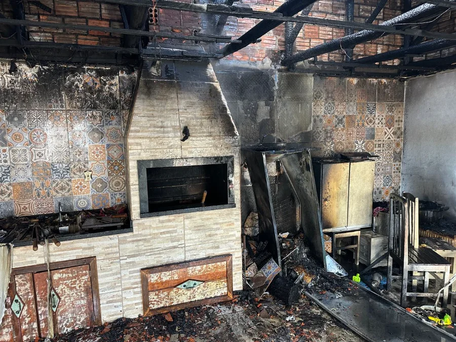 Interior de uma edícula em Forquilhinha após incêndio, mostrando a área da churrasqueira de alvenaria e móveis completamente carbonizados, com o telhado e as vigas de madeira destruídos.