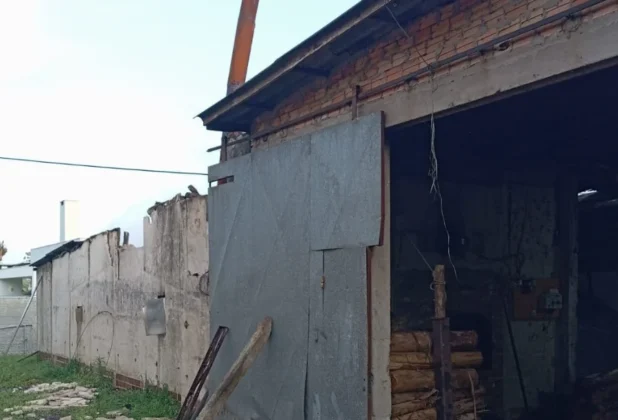 ista lateral da estufa de fumo após o incêndio, mostrando danos estruturais. A parede lateral esquerda está destruída e o telhado parcialmente desabou. Um grande portão de correr metálico está aberto, revelando o interior com vigas e sacos de fumo.