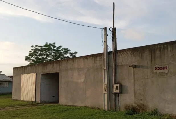 Estufa de fumo após o incêndio, vista do exterior. O edifício é de alvenaria e tem um portão de metal fechado à esquerda e um poste de energia elétrica com fiação na lateral da parede. O céu está claro e grama verde aparece na parte inferior.