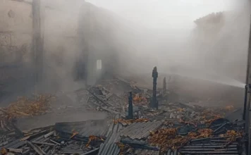 Incêndio atinge estufa de fumo no bairro Santa Fé, em Sombrio nterior da estufa de fumo em chamas. Fumaça densa preenche o ambiente. Escombros carbonizados, telhas metálicas destruídas e fardos de fumo queimados ou molhados estão espalhados pelo chão. Um jato de água, provavelmente de um bombeiro, é visível no canto superior direito.