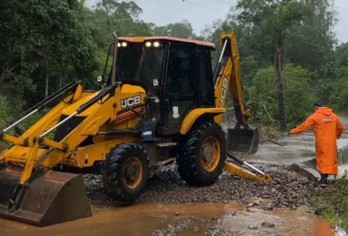 Jacinto Machado registra maior volume de chuvas de SC Retroescavadeira (máquina JCB) em estrada de chão alagada, com um funcionário da Defesa Civil ou Obras (de capa laranja) inspecionando danos, após Jacinto Machado registrar o maior volume de chuvas de Santa Catarina.
