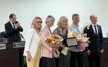 Luciane Ceretta recebe título de Cidadã Honorária de Turvo Luciane Ceretta recebendo flores e placa de homenagem de um grupo de pessoas em evento formal.