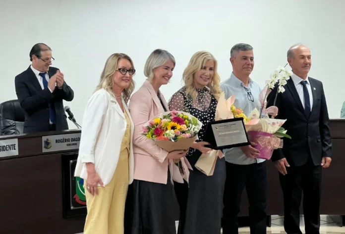Luciane Ceretta recebe título de Cidadã Honorária de Turvo Luciane Ceretta recebendo flores e placa de homenagem de um grupo de pessoas em evento formal.