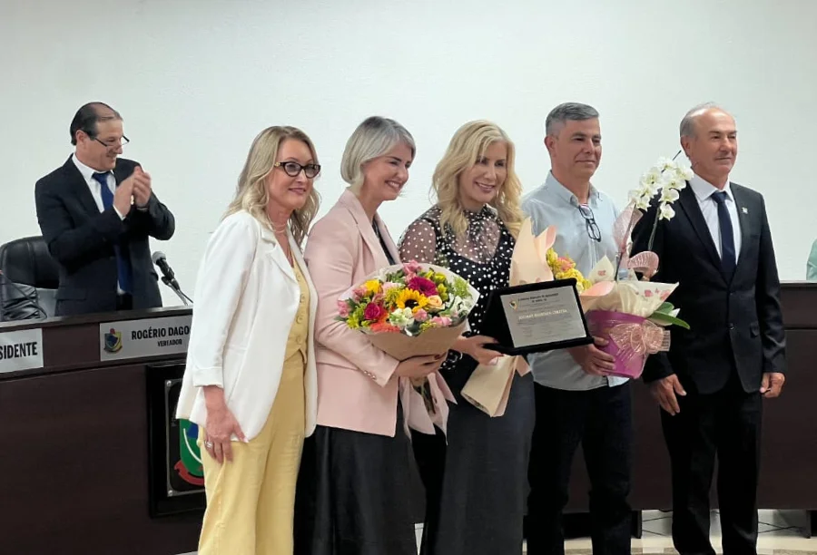 Luciane Ceretta recebendo flores e placa de homenagem de um grupo de pessoas em evento formal.