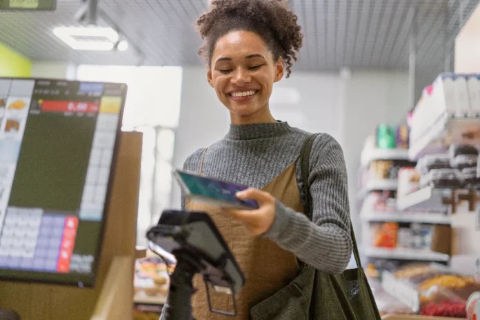 Mercado financeiro reduz previsão da inflação para 4,55% no Brasil Mulher sorrindo, pagando uma compra com o celular em um caixa (imagem ilustrativa), representando a confiança do consumidor em meio à redução da previsão da inflação (IPCA) para 4,55%.