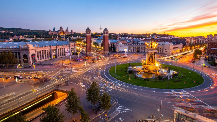 Vista noturna da Plaça d'Espanya em Barcelona, Espanha, com rastros de luzes de carros na rotatória, ilustrando o conceito de cidade inteligente e o foco da missão da FIESC em tecnologia urbana.