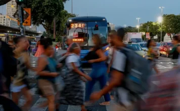 Mulheres ganham 15,8% menos que homens no mercado Pessoas atravessando uma rua movimentada no centro de uma cidade, com rastros de movimento (longa exposição) e um ônibus ao fundo, simbolizando a dinâmica do mercado de trabalho brasileiro e a desigualdade salarial de gênero (mulheres ganham 15,8% menos que homens).