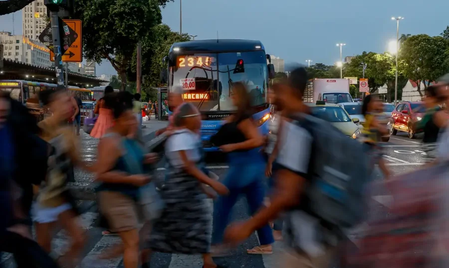 Pessoas atravessando uma rua movimentada no centro de uma cidade, com rastros de movimento (longa exposição) e um ônibus ao fundo, simbolizando a dinâmica do mercado de trabalho brasileiro e a desigualdade salarial de gênero (mulheres ganham 15,8% menos que homens).