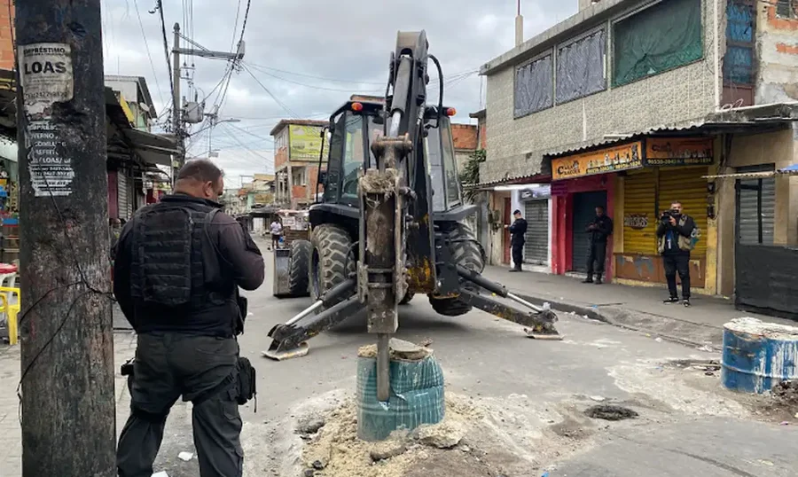 PL mira quem usa barricada para cometer ou ocultar outros crimes Policial de costas observa uma retroescavadeira removendo uma barricada em uma rua estreita de uma comunidade urbana.