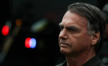 PL suspende funções e salário do ex-presidente Bolsonaro Ex-presidente Jair Bolsonaro em plano detalhe, com expressão séria; luzes de emergência de veículos policiais, embaçadas, aparecem no fundo.