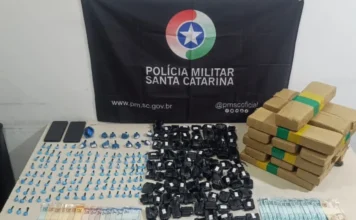 Passo de Torres: Operação da PM prende dupla por tráfico Mesa de apreensão com pacotes de maconha (tabletes e fracionada), cocaína, celulares e notas de dinheiro, dispostos em frente à bandeira da Polícia Militar de Santa Catarina.