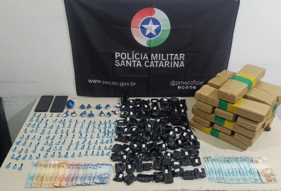 Passo de Torres Operação da PM prende dupla por tráfico Mesa de apreensão com pacotes de maconha (tabletes e fracionada), cocaína, celulares e notas de dinheiro, dispostos em frente à bandeira da Polícia Militar de Santa Catarina.