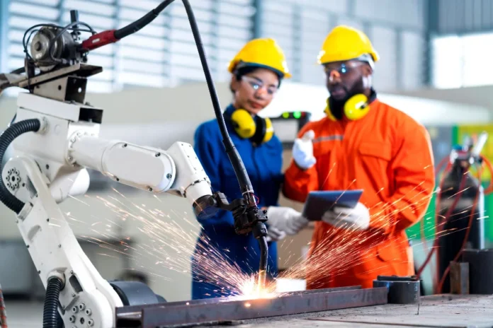 Produção industrial de SC cresce 3,1% no acumulado Trabalhadores com capacetes e abafadores (EPIs) supervisionando um braço robótico que realiza soldagem em uma fábrica, simbolizando o avanço de 3,1% na produção industrial de Santa Catarina e o foco em setores de alto valor agregado.