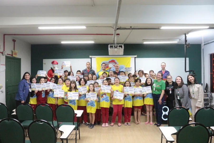 Cerca de 50 alunos do Colégio Unesc (Criciúma) com instrutores e mascote do Corpo de Bombeiros, exibindo os certificados de formatura do Programa Golfinho, que visa capacitar crianças em prevenção de acidentes aquáticos antes do verão.