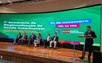 SES/SC debate gestão para fortalecer redes de saúde Auditório da SES com painel digital exibindo o tema "1º Seminário de Regionalização da Saúde Catarinense", com sete palestrantes sentados no palco e um orador no púlpito.