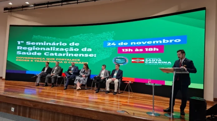 SES SC debate gestão para fortalecer redes de saúde Auditório da SES com painel digital exibindo o tema "1º Seminário de Regionalização da Saúde Catarinense", com sete palestrantes sentados no palco e um orador no púlpito.