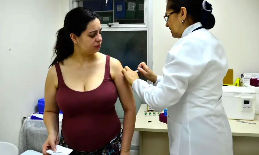 Profissional de saúde aplicando uma vacina no braço de uma mulher grávida, em uma Unidade Básica de Saúde.