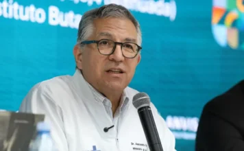 SUS vai ampliar em 30% aproveitamento de plasma O ministro da Saúde, Alexandre Padilha, em um close durante uma coletiva de imprensa, usando óculos e um jaleco médico branco. Ele está falando em um microfone em frente a um fundo turquesa.